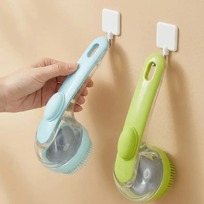 A hand holds a light blue Calivo™ Pet Bath & Grooming Brush with a shampoo dispenser, while an identical lime green one hangs on a hook beside it.